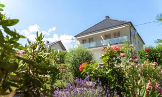 A beautiful house surrounded by colorful flowers and green bushes. The sky is clear and blue.