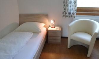 A simple bedroom with a single bed and a white seating area. Next to the bed, there is a bedside table with a lamp.