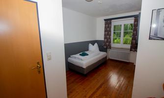 A simple room with a wooden floor and a bed. The windows offer a view of a green surroundings.