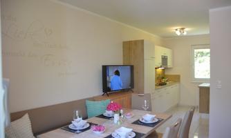 A bright, modern kitchen with a dining area. The table is set and a television is visible in the room.