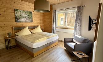 A cozy bedroom with a double bed and wooden walls. A small armchair and a table complete the furnishings.