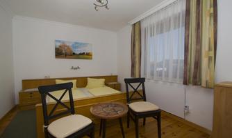 A simple bedroom with a bed and two chairs.  
Light-filled windows and a picture on the wall create an inviting atmosphere.