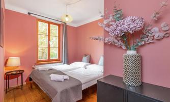 A cozy bedroom with a pink wall. There is a double bed, a small table, and a stylish vase with flowers.
