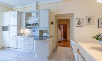 A bright and modern kitchen with white furniture and elegant tiles. In the background, a dining table and a passage to another room are visible.
