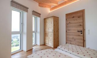 A modern bedroom with wooden floor and large windows. The room has a wooden wardrobe and two beds with patterned bedding.