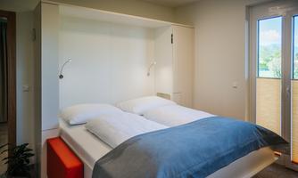 A modern bedroom with a double bed and a large window. The decor is bright and minimalist, featuring a blue blanket and an orange bench.