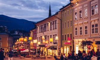 A picturesque city square at twilight with illuminated buildings and cozy seating. People are sitting outside and enjoying the atmosphere.