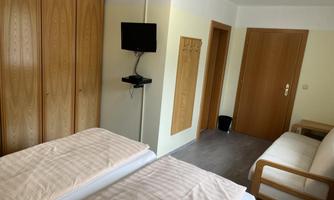 A simple room with two single beds, a television, and a sofa. The walls are painted light and there is a wardrobe as well as a door to another room.