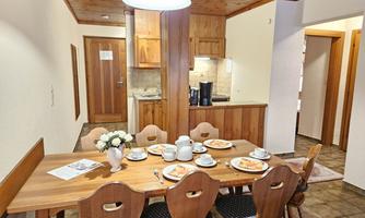 An inviting dining area with a large wooden table and chairs. On the table, there are dishes and breakfast prepared, and the kitchen in the background is designed in a modern style.
