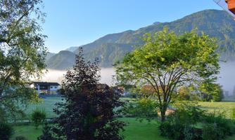 A picturesque landscape with mountains in the background and trees in the foreground. The sky is clear and blue, while fog hovers over the valley.