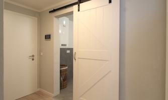 A modern interior with a white sliding door and a light wall color. In the background, a well-equipped bathroom can be seen.