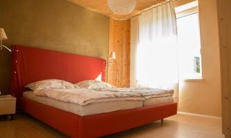 A cozy bedroom with a red bed and bright cushions. The room is well-lit and has a large window with curtains.
