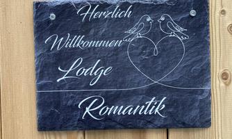 A welcome sign made of black stone, displaying the words "Warm Welcome" and "Lodge Romance." Two birds are depicted in a heart motif.