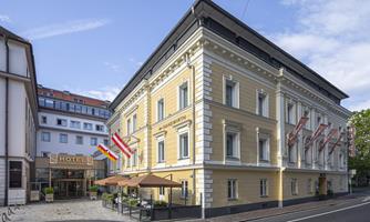 An elegant hotel building with a yellow facade and flags. Outdoor furniture is located in front of the entrance and the street area is well-maintained.