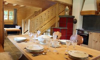 A rustic dining room with a long wooden table, lovingly set for a feast. In the background, there is a staircase and a modern kitchenette.