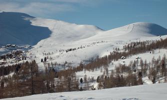 A snowy landscape with gentle hills and mountains in the background. The sky is clear and blue.