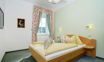 A cozy bedroom with a wooden bed and colorful cushions. Bright walls and a window with curtains create a pleasant atmosphere.