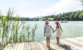 Two girls are standing at the edge of a clear lake, holding hands. In the background, there are lush trees and gentle hills.