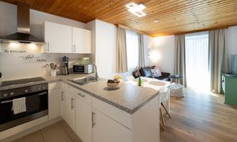 A modern kitchen with an open living area. The space is bright and inviting, with a wooden ceiling and large windows.