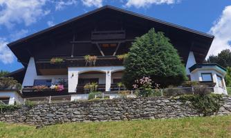 A large, traditional wooden house with flower balconies and a green hedge. The sky is blue with some clouds.