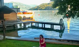 A woman is sitting on a meadow by the water, surrounded by mountains. She has spread out a blanket and is playing with singing bowls.