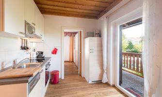 A modern kitchen with wooden flooring and light furniture. The windows offer a view of a balcony and plenty of daylight.