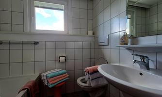 A modern bathroom with white tiles, a sink, a toilet, and a bathtub. Colorful towels are neatly stacked, and a window provides daylight.
