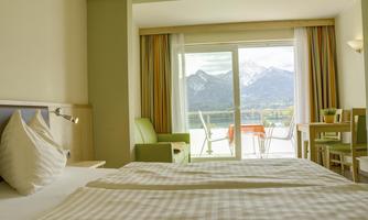 A bright hotel room with a view of the mountains. Outside, there is a table with chairs on the balcony.