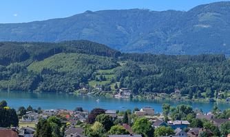 A picturesque view of a lake surrounded by green mountains and a clear blue sky. In the foreground, small houses are visible.