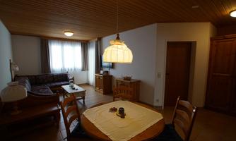 A cozy living space with a dining table and chairs. In the background, there is a couch and a window with curtains.