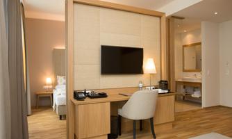 A modern hotel room with a flat-screen TV, a desk, and a cozy seating area. The warm wooden floor and soft color tones create an inviting atmosphere.