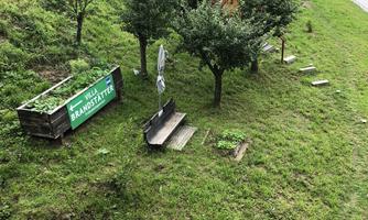 A green meadow with fruit trees and small huts. In the foreground are plant boxes and a path.