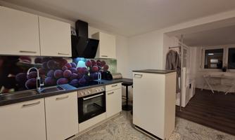 A modern kitchen with white cabinets and a view of purple grapes in the wall decoration. In the background, a table and chairs can be seen.