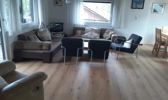 A cozy living room with a sofa and several chairs.  
Large windows let in plenty of light and the wooden floor creates a warm atmosphere.