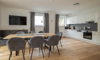 A modernly furnished living space with a dining area and an open kitchen. The decor is bright and friendly with hardwood floors and elegant chairs.