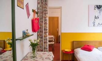 A cozy room with two beds, colorful walls, and a small table. A bouquet of flowers is on the table and a door leads to another room.