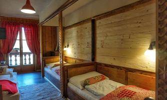 A cozy room with two wooden beds and warm colors. Large windows with red curtains offer a view outside.