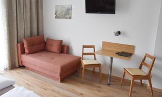 A simply furnished room with a sofa and a table with two chairs. A television hangs on the wall, and there is a window with curtains.