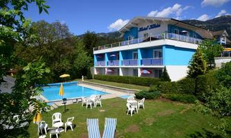 A beautiful hotel with a blue facade and a swimming pool. The garden is green and features lounge chairs as well as sun umbrellas.
