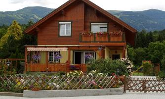 A charming wooden house with many flowers on the balcony and in the garden. In the background, green mountains can be seen.