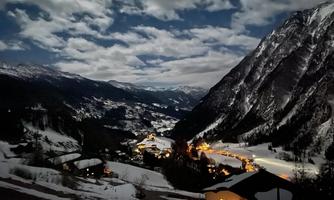 A magnificent mountain landscape at night, illuminated by cozy lights in the houses. The sky is adorned with clouds and the snow-covered hills are clearly visible.