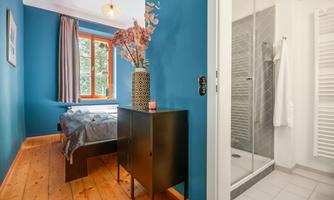 A cozy room with blue walls and wooden flooring. In the background, there is a bathroom with a modern shower visible.