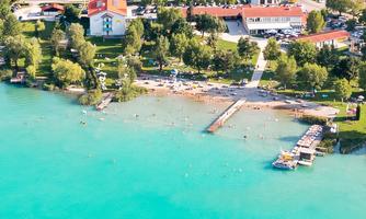 A beautiful lake with clear, turquoise water and a beach. Along the shore, there are trees and some buildings.