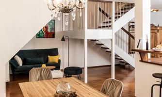 A modern dining room with a large wooden table and elegant chairs. A stylish lamp hangs from the ceiling and a staircase leads to the upper area.