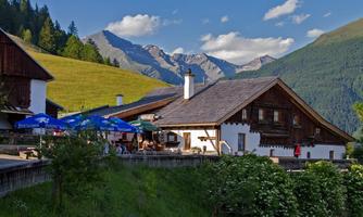 A traditional mountain hut surrounded by green meadows and mountains. In the foreground, you can see several umbrellas and guests enjoying the view.