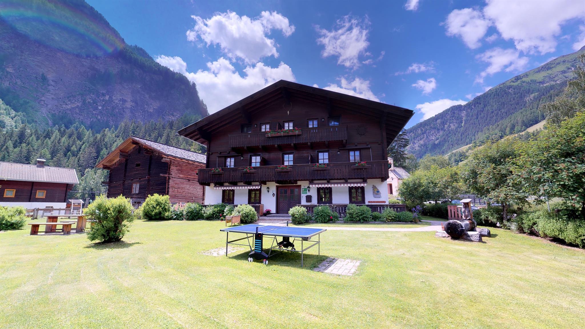 A charming wooden house surrounded by a green meadow and mountains. The sky is blue with a few clouds and there is a table tennis court in the foreground.