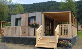 A modern wooden house with a porch and large windows. It is located in a natural environment, surrounded by trees and hills.