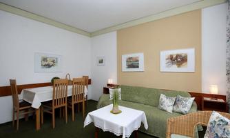 A cozy living room with a green sofa and a small table. In the background, there are pictures on the walls and a dining table with chairs.