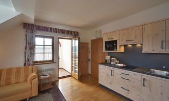 A bright kitchen with wooden cabinets and modern appliances. In the background, there is a living room with a sofa and a window that leads outside.