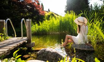 A woman sits relaxed by the water. Green plants grow around her and the sun shines on the scene.